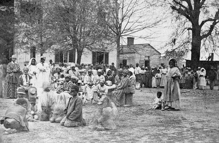 This historical black and white photograph depicts a large gathering of African American people, most likely emancipated slaves, at a contraband camp during the Civil War era. Men, women, and children are seen in various attire, some standing, some seated on the ground. In the foreground, a few individuals appear to be engaged in activities, possibly cooking, as there is smoke rising from a spot on the ground. There are two main structures in the background, simple wooden buildings that might have been used for living quarters or other camp functions. Trees are visible around the area, indicating a rural setting. The image captures a moment of daily life in a community that formed in the wake of emancipation, highlighting the resilience and adaptability of those seeking freedom and a new life.