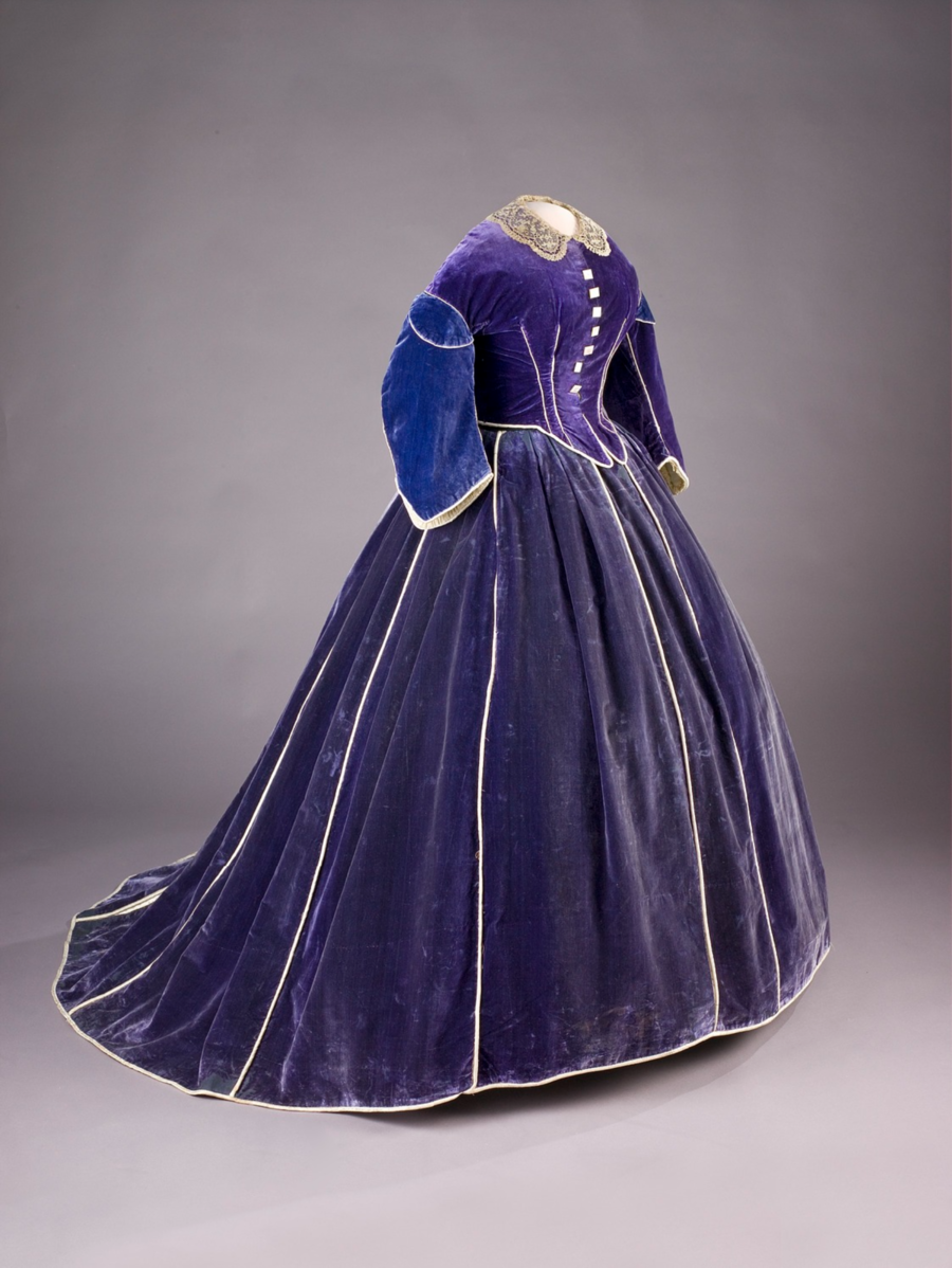 A portrait-oriented photo of an elegant, royal blue velvet period dress on a mannequin against a neutral gray background. This mid-19th century gown features a fitted bodice with a row of buttons and white lace collar, long, curved sleeves, and a full, bell-shaped skirt typical of the era. The dress is adorned with white piping along the seams and edges, enhancing its luxurious and formal appearance. The skirt's broad, smooth expanse and the velvet's plush texture reflect the light differently at various angles, indicating the high quality of the material and craftsmanship. Such a dress would have been worn by a woman of status or for a significant occasion, potentially representing the fashion of a first lady or a prominent female figure of that time.